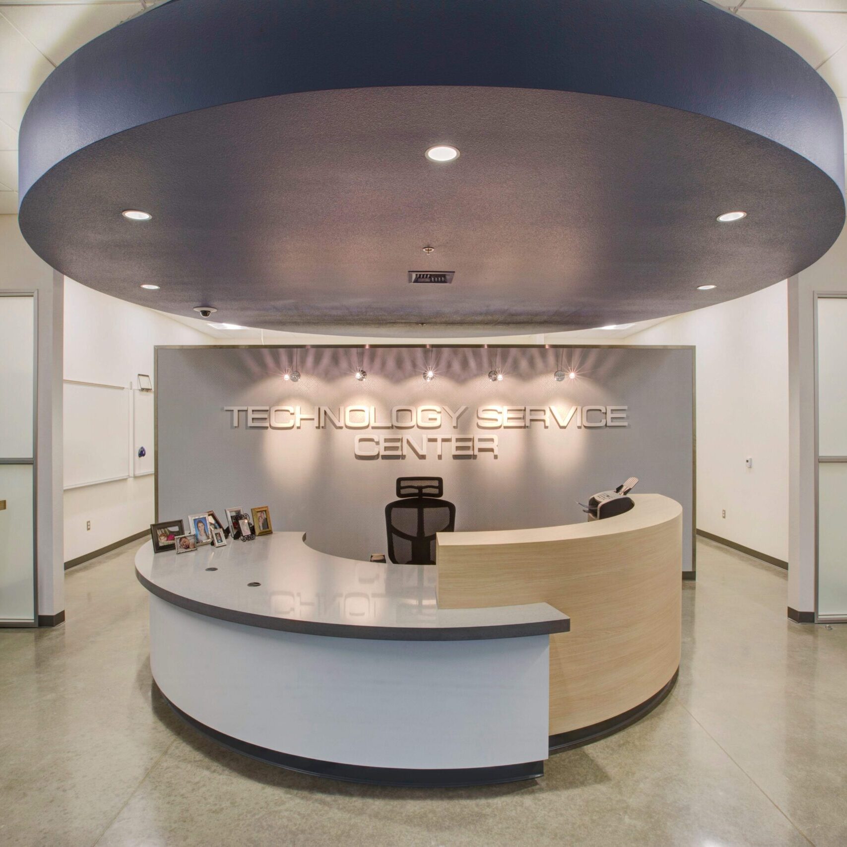 A centered, interior shot of a modern reception area. The main feature is a curved, two-toned reception desk with a light wood front and a gray top. Behind the desk, a large gray wall has three-dimensional letters spelling out "TECHNOLOGY SERVICE CENTER." Above the desk, a large, dark blue circular fixture is suspended from the ceiling with recessed lighting. The floor is a polished concrete surface. To the left, there is a small counter with a computer and stool, and to the right, a wooden door is visible. The lighting is bright and the space appears clean and modern.