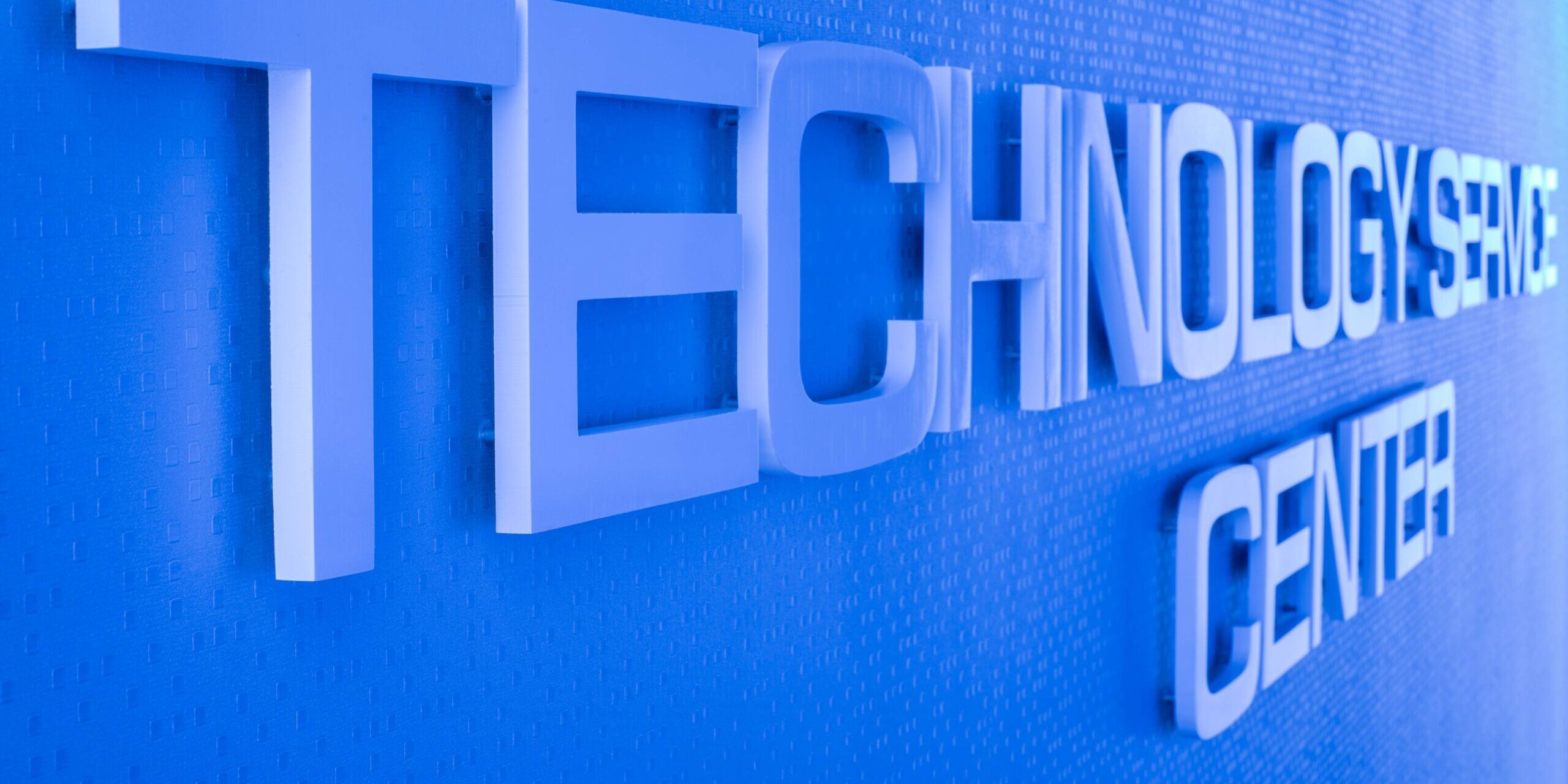 A slightly angled, close-up shot of a sign on a wall inside the building. The words "TECHNOLOGY CENTER" are spelled out in large, three-dimensional, light-colored letters mounted on a vibrant blue wall with a subtle textured pattern. The lighting highlights the shadow and depth of the letters. The sign is designed with "TECHNOLOGY" on the top line and "CENTER" on the bottom line.