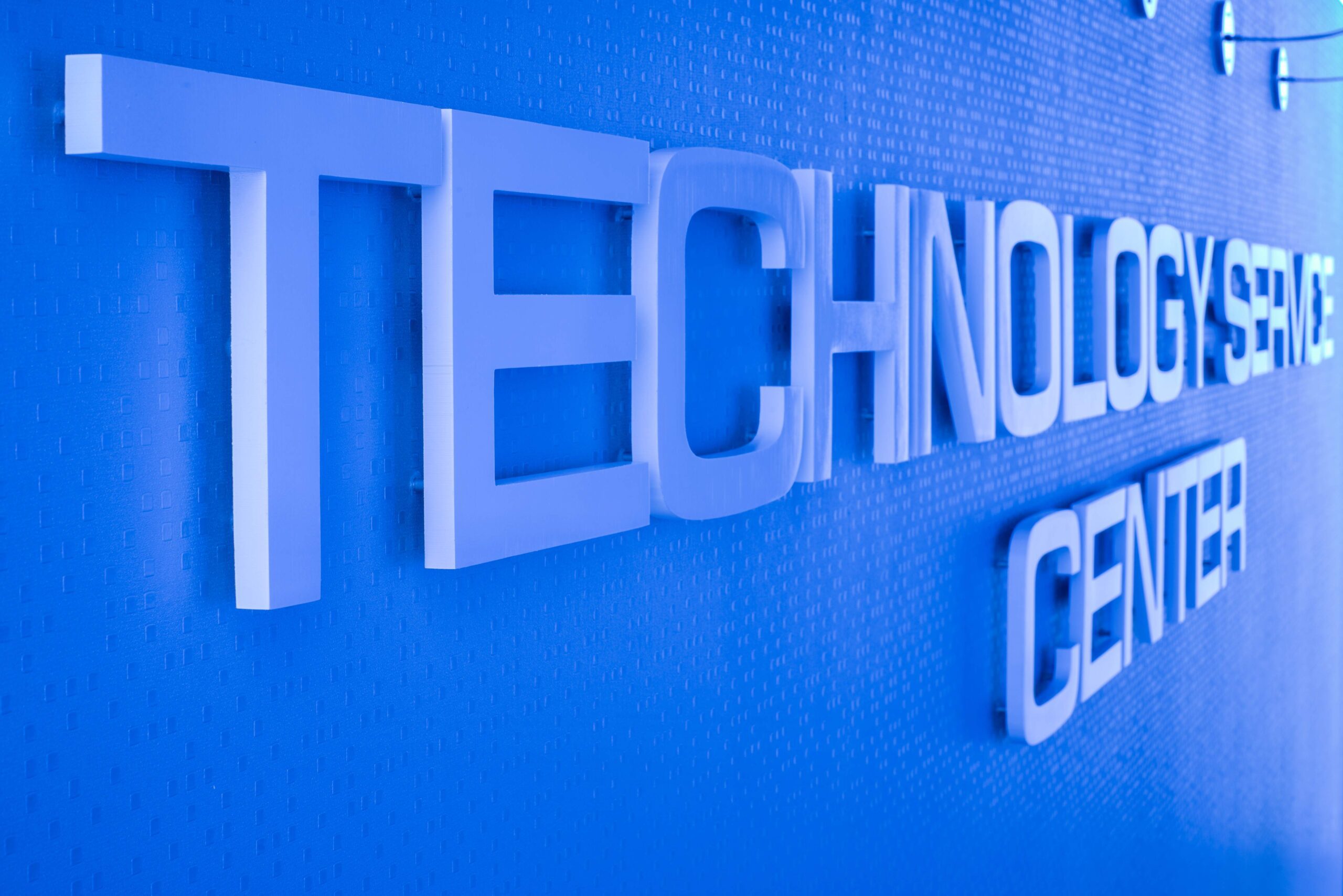 A slightly angled, close-up shot of a sign on a wall inside the building. The words "TECHNOLOGY CENTER" are spelled out in large, three-dimensional, light-colored letters mounted on a vibrant blue wall with a subtle textured pattern. The lighting highlights the shadow and depth of the letters. The sign is designed with "TECHNOLOGY" on the top line and "CENTER" on the bottom line.