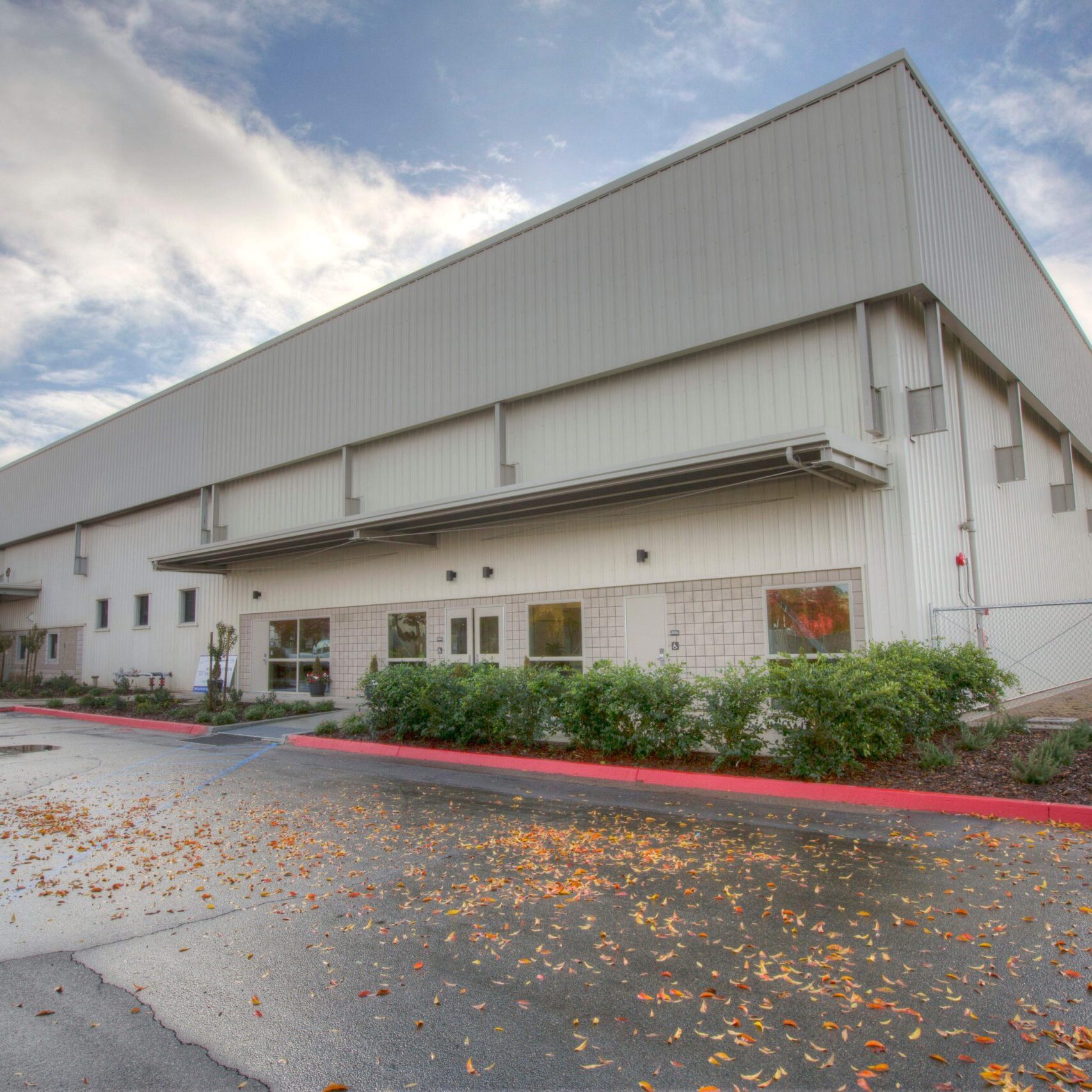 A wide-angle exterior shot of a large, modern, industrial-style building. The building has a long, gray metal-paneled facade with a lower section of white brick and large windows. A red curb lines the front of the building, and a paved parking lot with a few fallen leaves is in the foreground. The sky is partly cloudy, with dramatic clouds overhead. The building is long, with what appears to be a smaller fenced-in area to the right. A few trees with autumnal leaves are visible on the left side of the frame.