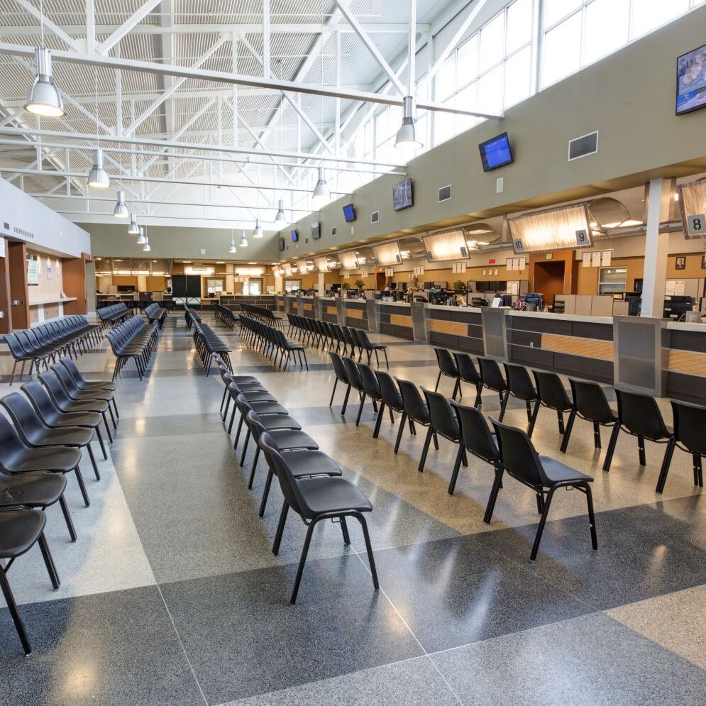 A wide-angle, indoor photo of a large, empty waiting room, likely a DMV or government office. Rows of black plastic chairs are arranged on a speckled gray and white tiled floor, facing a long counter with multiple service windows. The ceiling is high with exposed metal trusses and fluorescent lighting. Large windows are visible at the top of the walls, letting in natural light. A few monitors are mounted on the wall above the counters.