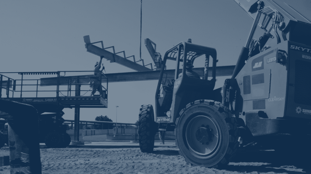A blue-toned photograph of a construction site. In the foreground, a large, green "SKYTRAK" telehandler is parked on a dirt lot. A person in a hardhat is in the driver's seat. In the background, a person in an orange safety vest is standing in a lift, assisting with the placement of a large steel beam that is being hoisted by a crane. The sky is a light blue.