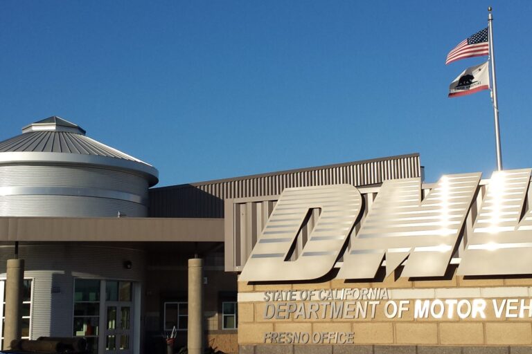 A close-up, outdoor shot of the front of a modern DMV building. The building has a large, metallic, stylized "DMV" sign. Below it, the text "STATE OF CALIFORNIA DEPARTMENT OF MOTOR VEHICLES FRESNO OFFICE" is visible. A circular, metallic section of the building with a dome roof is visible to the left, and a flagpole with the American and California state flags is visible to the right. The sky is a clear blue.