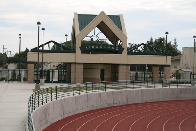 A medium-angle exterior shot of the entrance to a sports stadium at Kingsburg High School. The entrance is a two-story structure with a peaked green roof and a tan stone facade. The word "KINGSBURG" is visible in large letters above the archway. A concrete track with red lanes is in the foreground, curving to the right, with a black metal railing separating it from the paved concourse area. The sky is overcast. In the background, there are residential buildings and trees. The overall image captures the entrance to a well-maintained athletic facility.