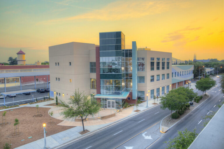 A professional, outdoor photo of a multi-story, modern building with tan and red facades and a prominent glass-enclosed section. The building is located next to a street with a few trees and streetlights, and a red brick building with a tower is visible in the background. The photo is taken at sunset, with a vibrant orange and yellow sky.