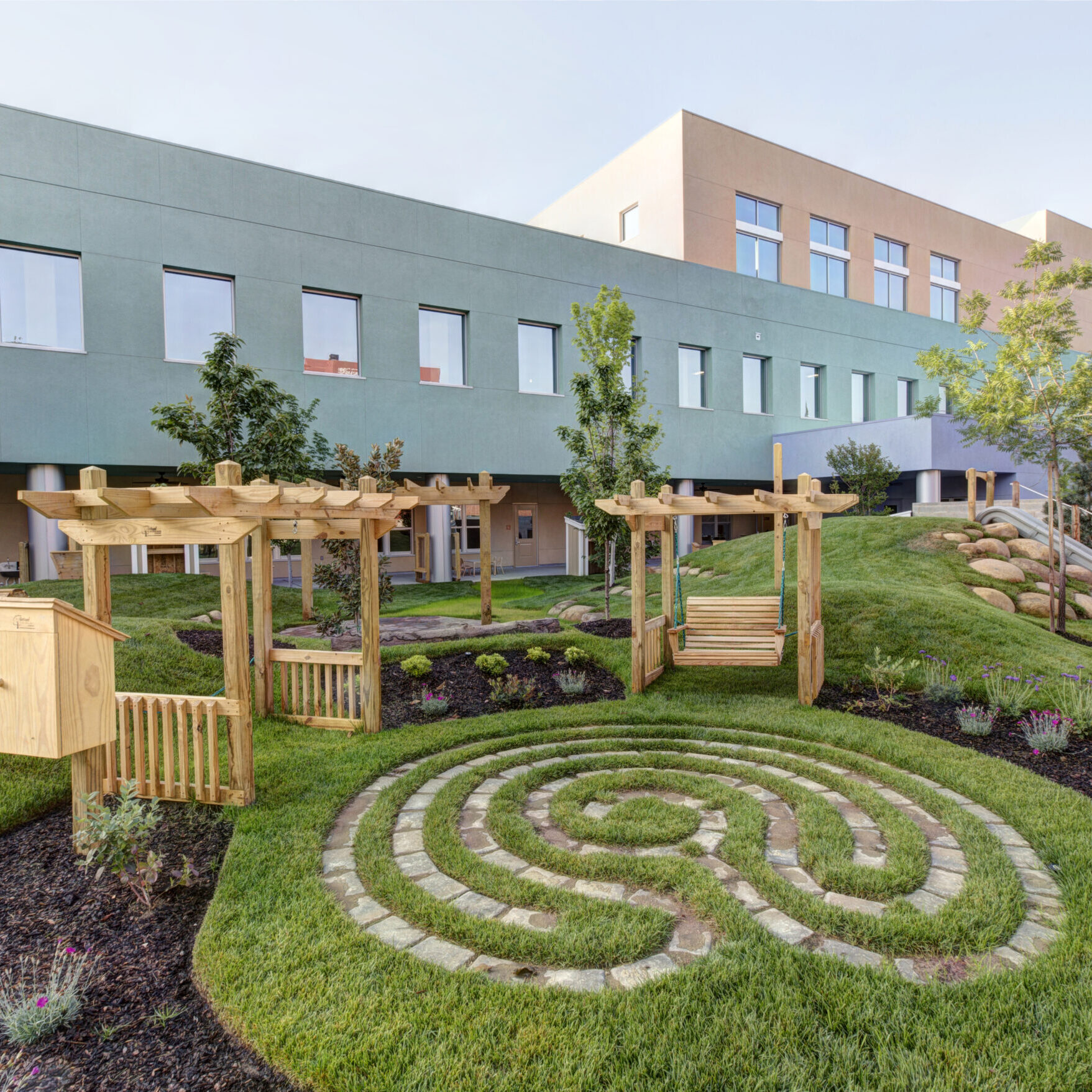 A wide, professional photo of a modern office building with a well-maintained outdoor garden and recreation area. The building has a light green and tan facade with rows of windows. The garden in the foreground features a grass labyrinth outlined with gray pavers, a wooden pergola with a swing, and a small slide built into a grassy hill. A few trees are planted throughout the area, and the ground is covered with grass and dark mulch.