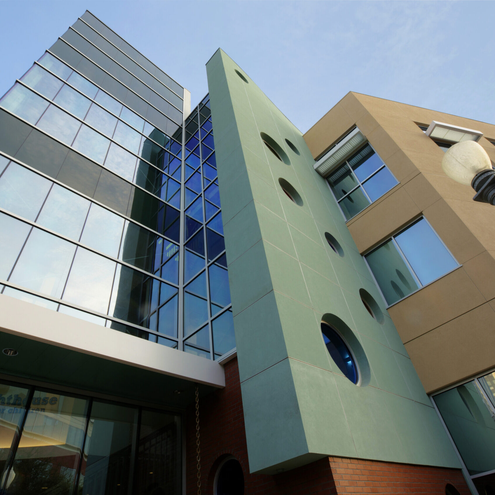 A dynamic, low-angle shot of a multi-story building with a unique, modern architectural design. The building has a tan-colored facade on the right and a glass facade on the left, reflecting the blue sky. In the center, a light green, triangular-shaped structure with circular windows rises between the two wings. A street lamp is visible on the right.