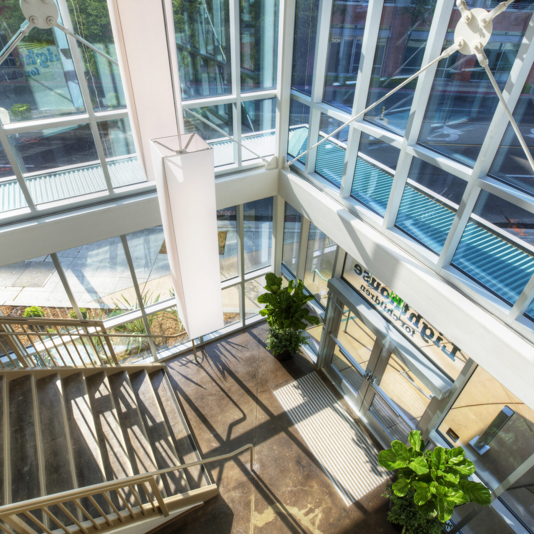 A top-down, indoor shot of a modern building lobby with a large glass facade. A staircase with a wooden handrail descends from the upper left. The floor is a polished concrete, and a large column is in the center. The glass walls and ceiling are supported by a metal frame, letting in a large amount of natural light. Two potted plants sit near the entrance, and the text on the glass doors is mirrored and reads "lighthouse".