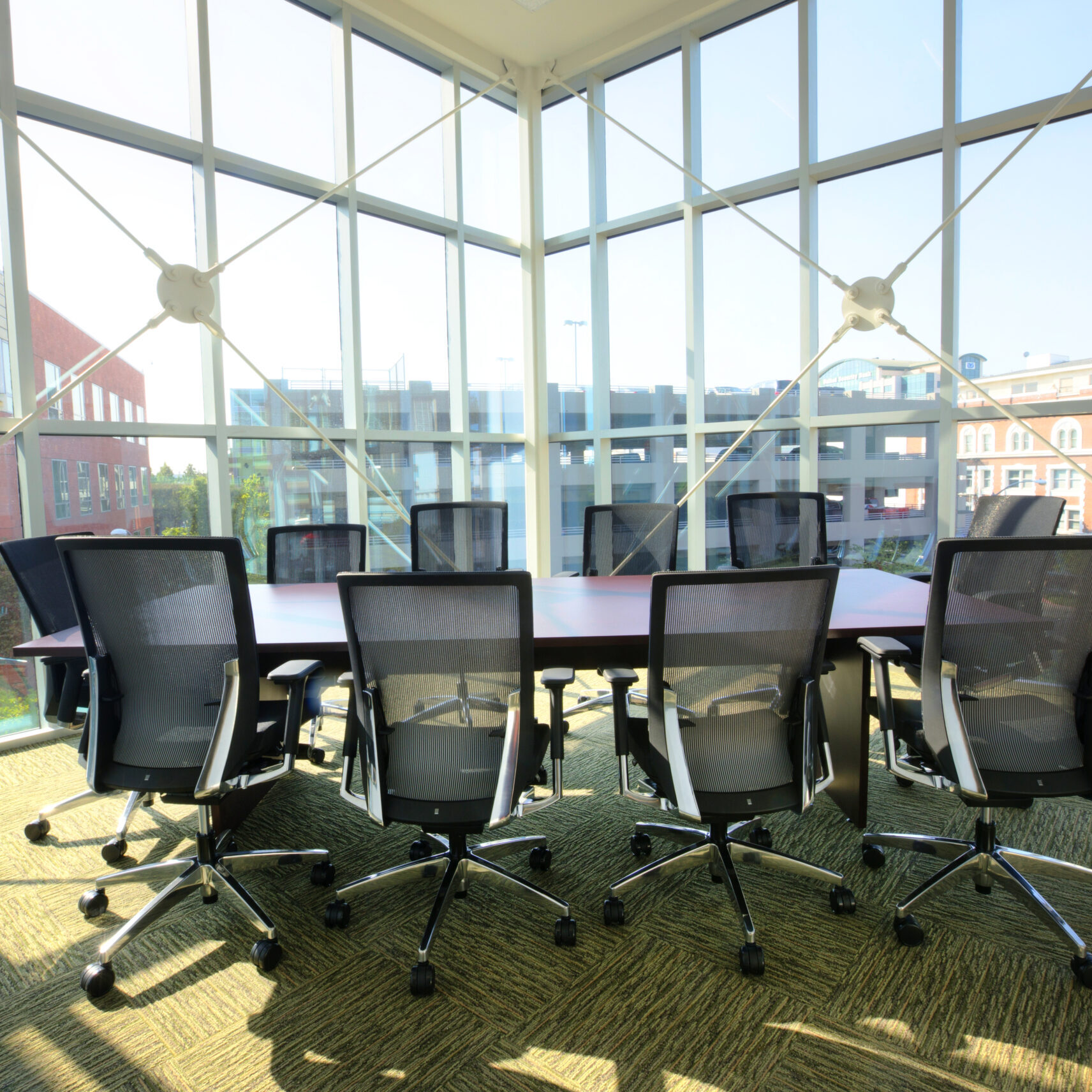 A professional, wide-angle indoor photo of a modern conference room. The room has a large table surrounded by black and gray chairs on a green carpet. The walls are made entirely of glass, offering a panoramic view of the buildings and cityscape outside. The glass is held together by a white metal frame.