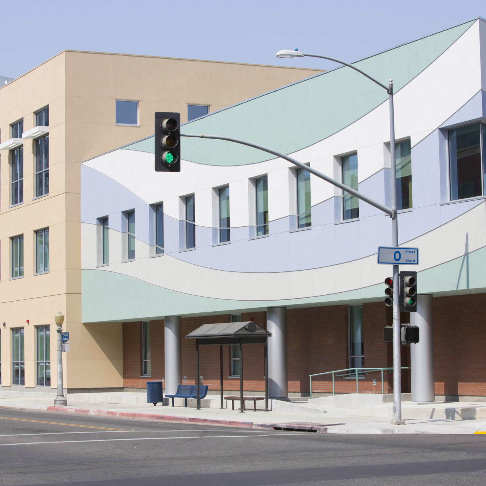 A wide-angle, outdoor shot of a modern, multi-story building. The building has a tan facade on the left and a front section with a white, light blue, and light green wave pattern. A street with traffic lights and a bus stop with a bench is in the foreground. In the background, a tall, older brick building is partially visible on the left.