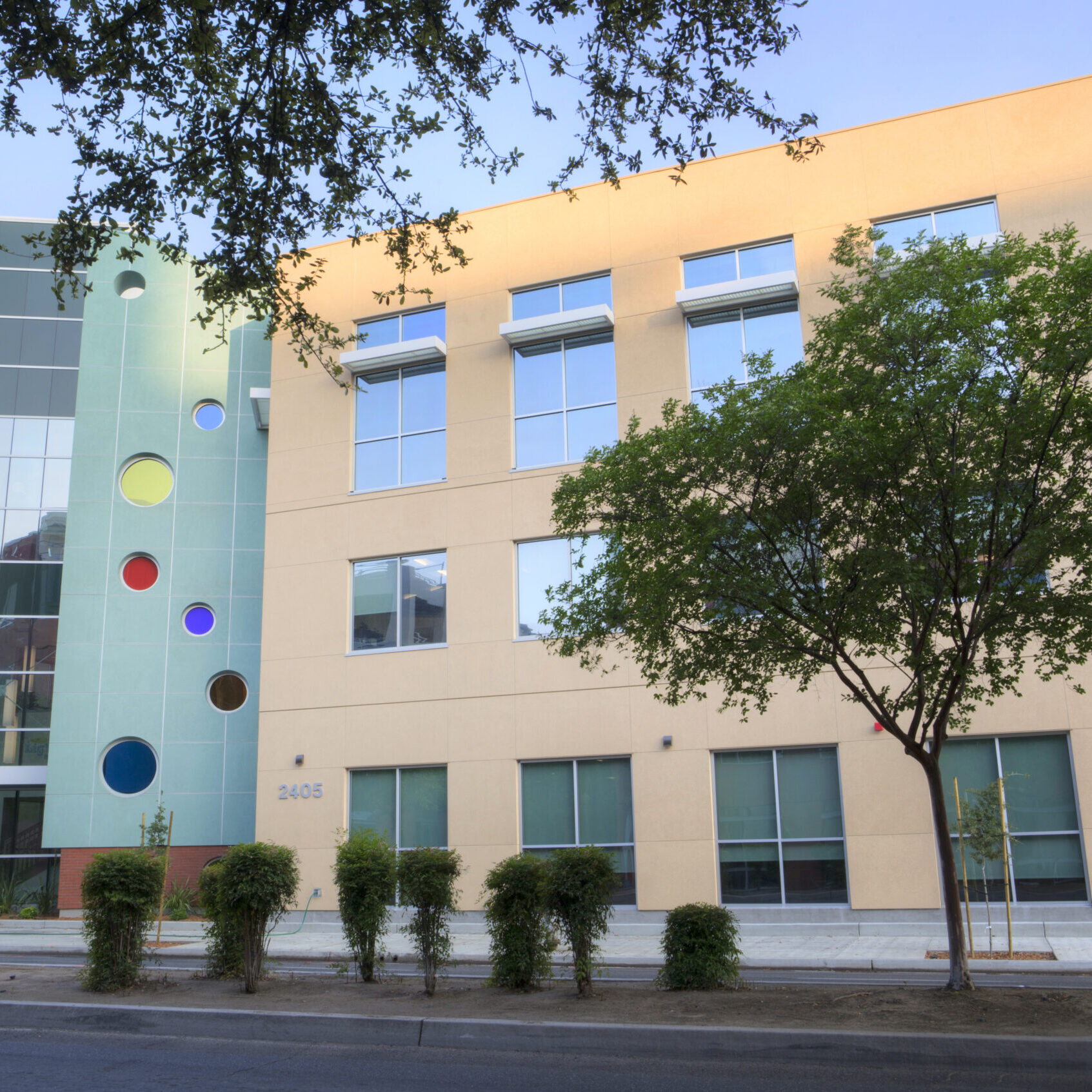 A professional, outdoor photo of a multi-story, modern building with a mix of architectural styles. The building has a light tan facade with large windows, and an address number "2405" is visible on the first floor. A distinctive light green section with circular windows of different colors is visible to the left. The foreground features a sidewalk, street, and a few trees. The sky is a clear blue.