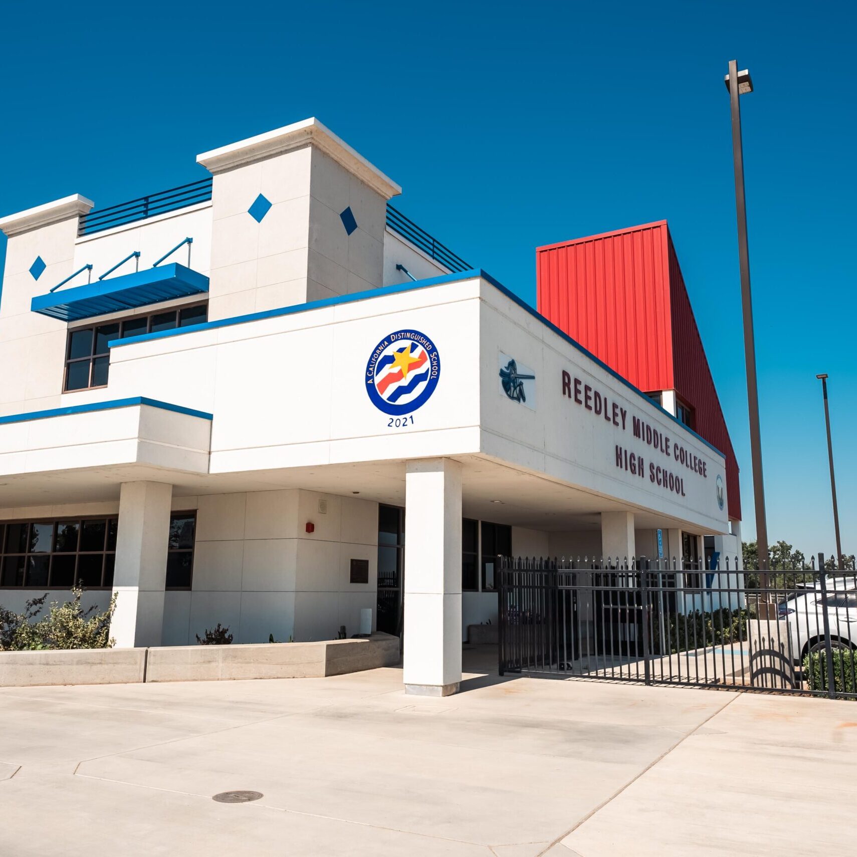A professional, wide-angle, outdoor photo of the front of a modern high school building. The building has a white stucco exterior with blue trim and a red metal roof section. The school name, "REEDLEY MIDDLE COLLEGE HIGH SCHOOL," is visible on the facade. A circular logo with a wave design and the year "2021" is also visible. The entrance is covered by a blue awning, and a black fence runs along the front of the building. The sky is a clear, bright blue.