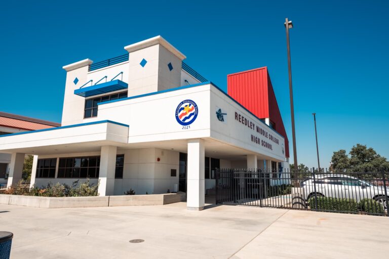 A professional, wide-angle, outdoor photo of the front of a modern high school building. The building has a white stucco exterior with blue trim and a red metal roof section. The school name, "REEDLEY MIDDLE COLLEGE HIGH SCHOOL," is visible on the facade. A circular logo with a wave design and the year "2021" is also visible. The entrance is covered by a blue awning, and a black fence runs along the front of the building. The sky is a clear, bright blue.