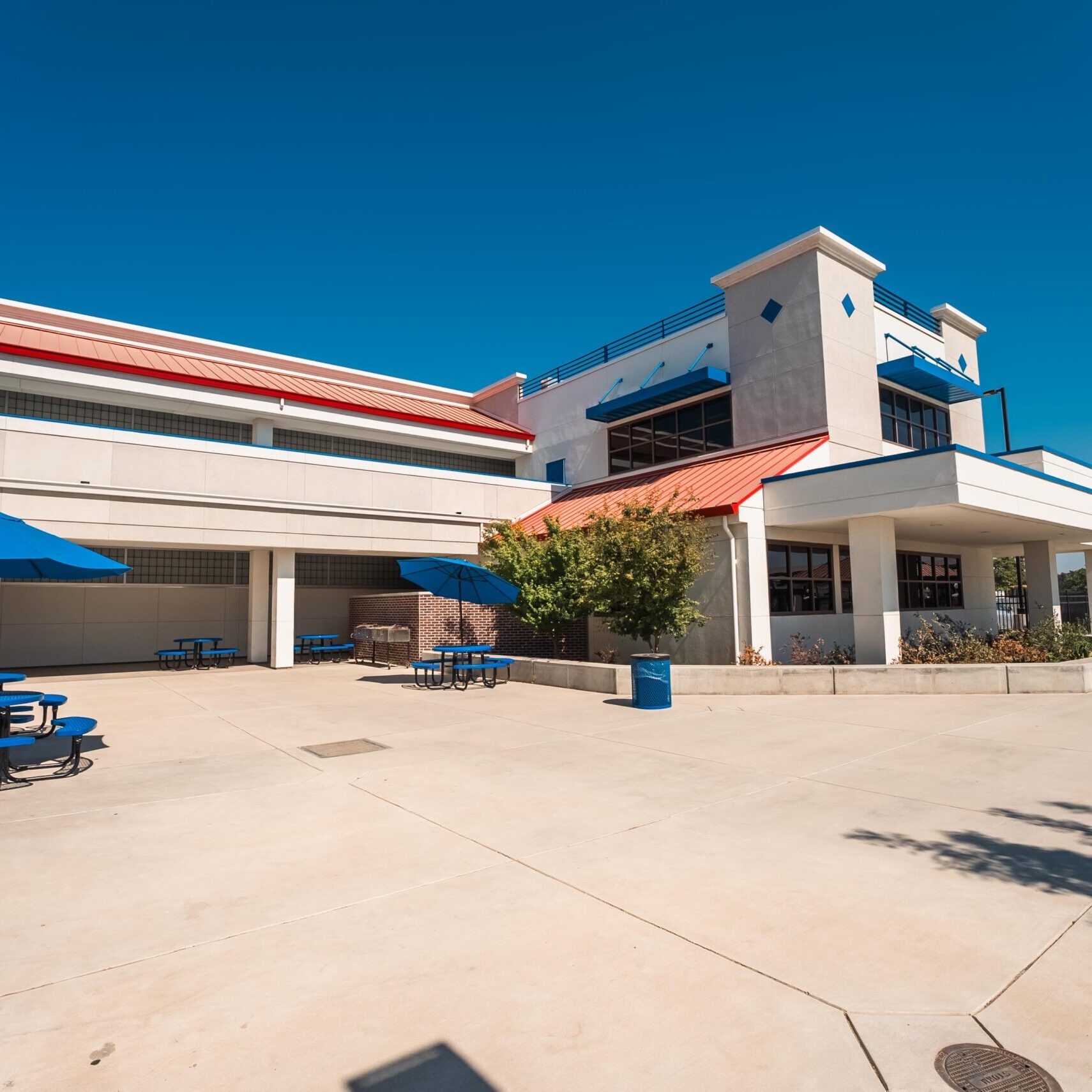A wide-angle, outdoor shot of a modern school campus courtyard. The courtyard has a light-colored paved surface, with several blue picnic tables and umbrellas. The building has a white and tan stucco exterior with blue trim and red metal roof sections. The sky is a clear, bright blue.