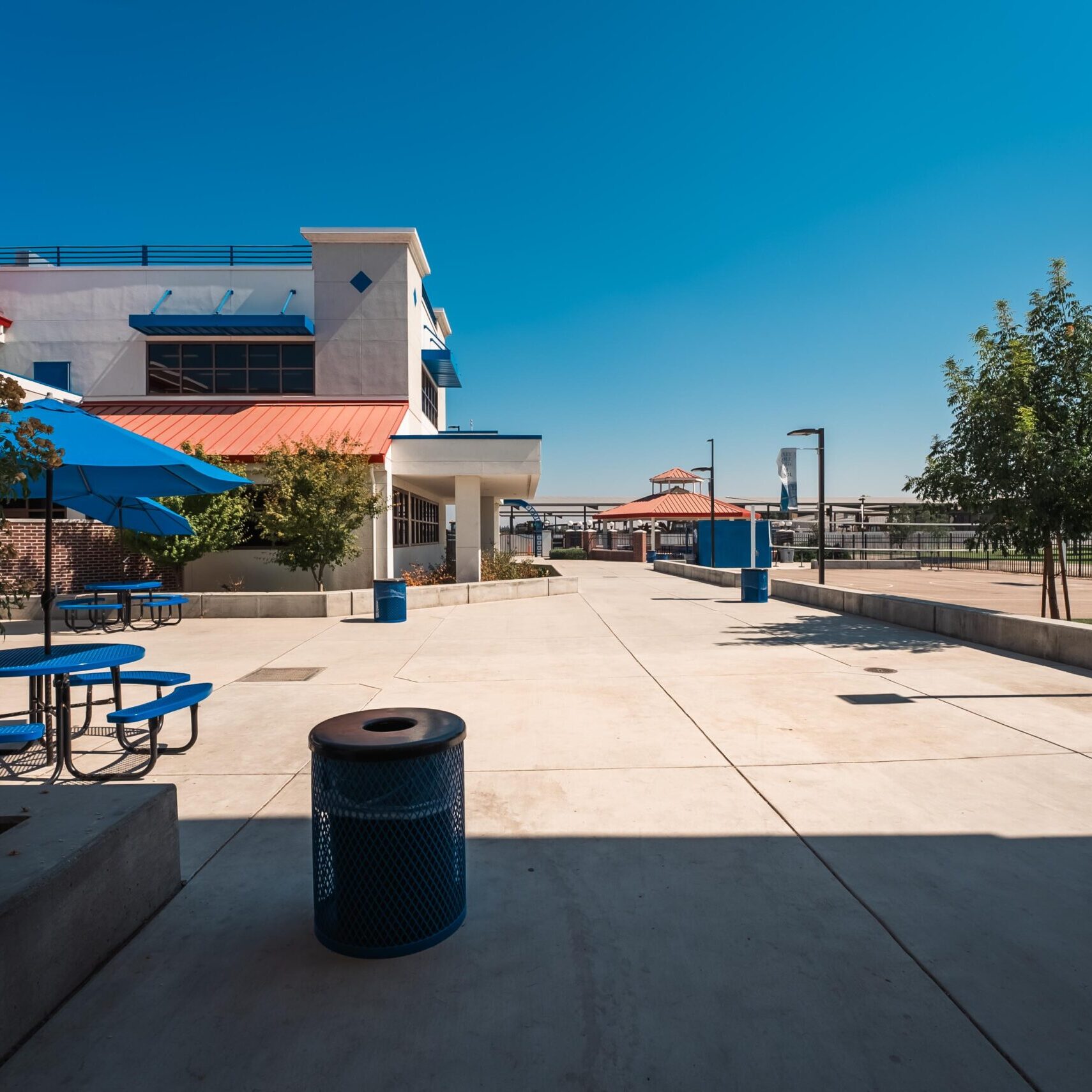 A wide-angle, outdoor shot of a modern school campus courtyard. The courtyard has a light-colored paved surface, with blue trash cans, and a few trees and bushes. To the left are blue picnic tables and umbrellas. A banner with the school name, "REEDLEY MIDDLE COLLEGE HIGH SCHOOL," is visible on a light post to the right. The buildings have white and red roofs, and the sky is a clear, bright blue.