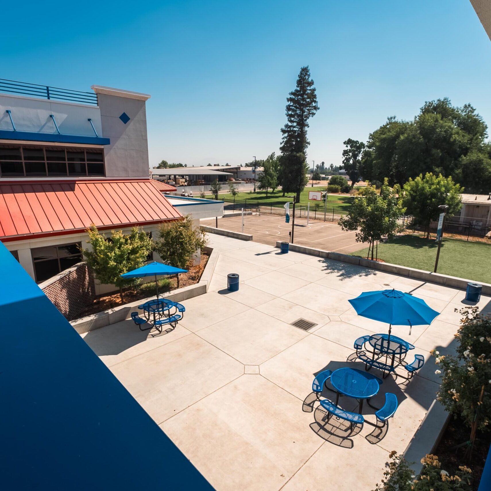An aerial, top-down shot of a school courtyard on a clear day. The courtyard has a light-colored paved surface with two sets of blue picnic tables and matching umbrellas. A large red-roofed section of a building is visible to the left. The photo is taken from a second-story balcony with a blue railing, looking down on the courtyard and a grassy area with a sports court in the distance. The sky is a clear, bright blue.