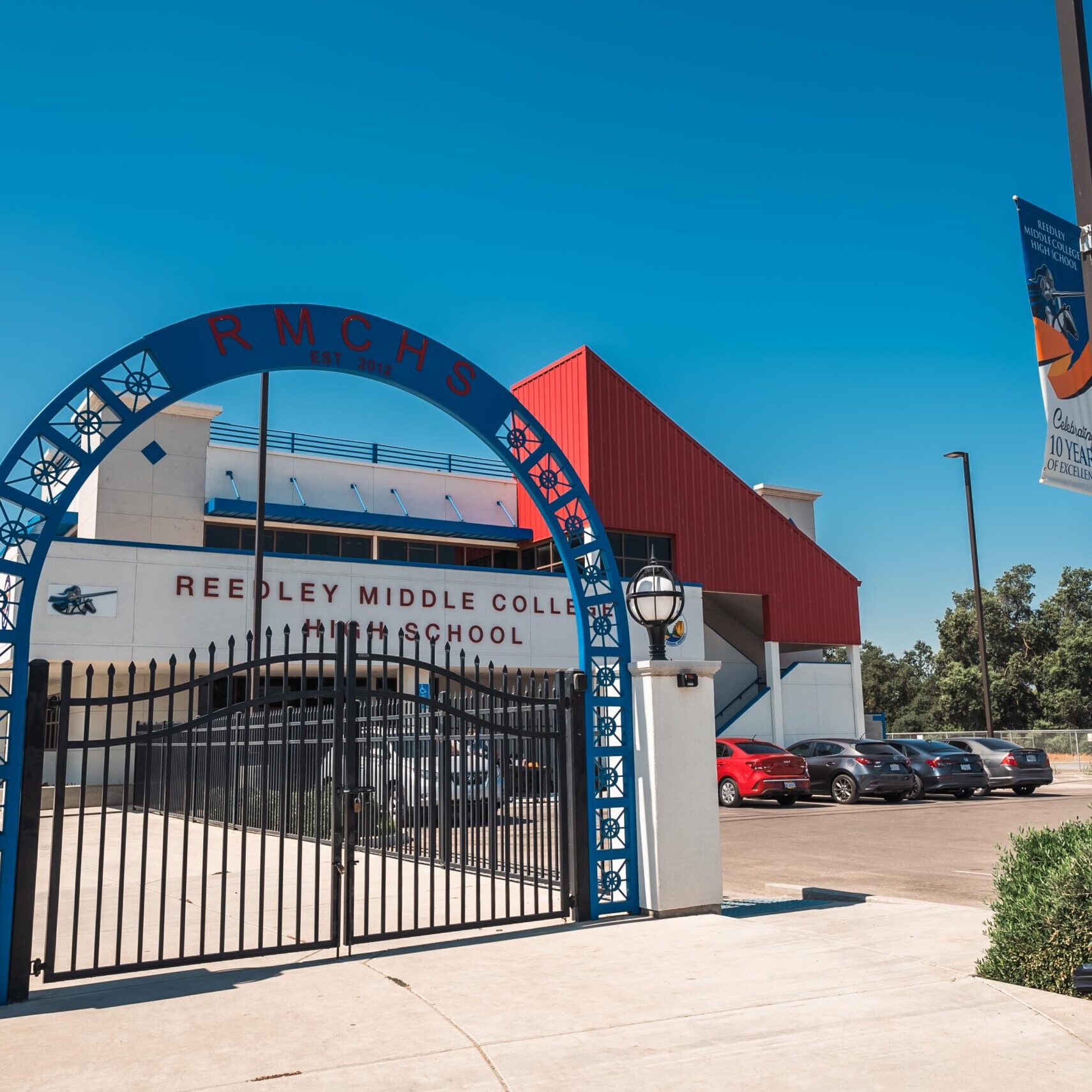 A wide, professional, outdoor shot of the entrance to a high school. The main gate is black with a large, arched top featuring a blue and white decorative design with the letters "RMCHS." The school building, with its white and red facade, is visible behind the gate. The name "REEDLEY MIDDLE COLLEGE HIGH SCHOOL" is on the front of the building. A banner is visible on a light post to the right, and a parking lot with cars is visible to the right of the building. The sky is a clear, bright blue.