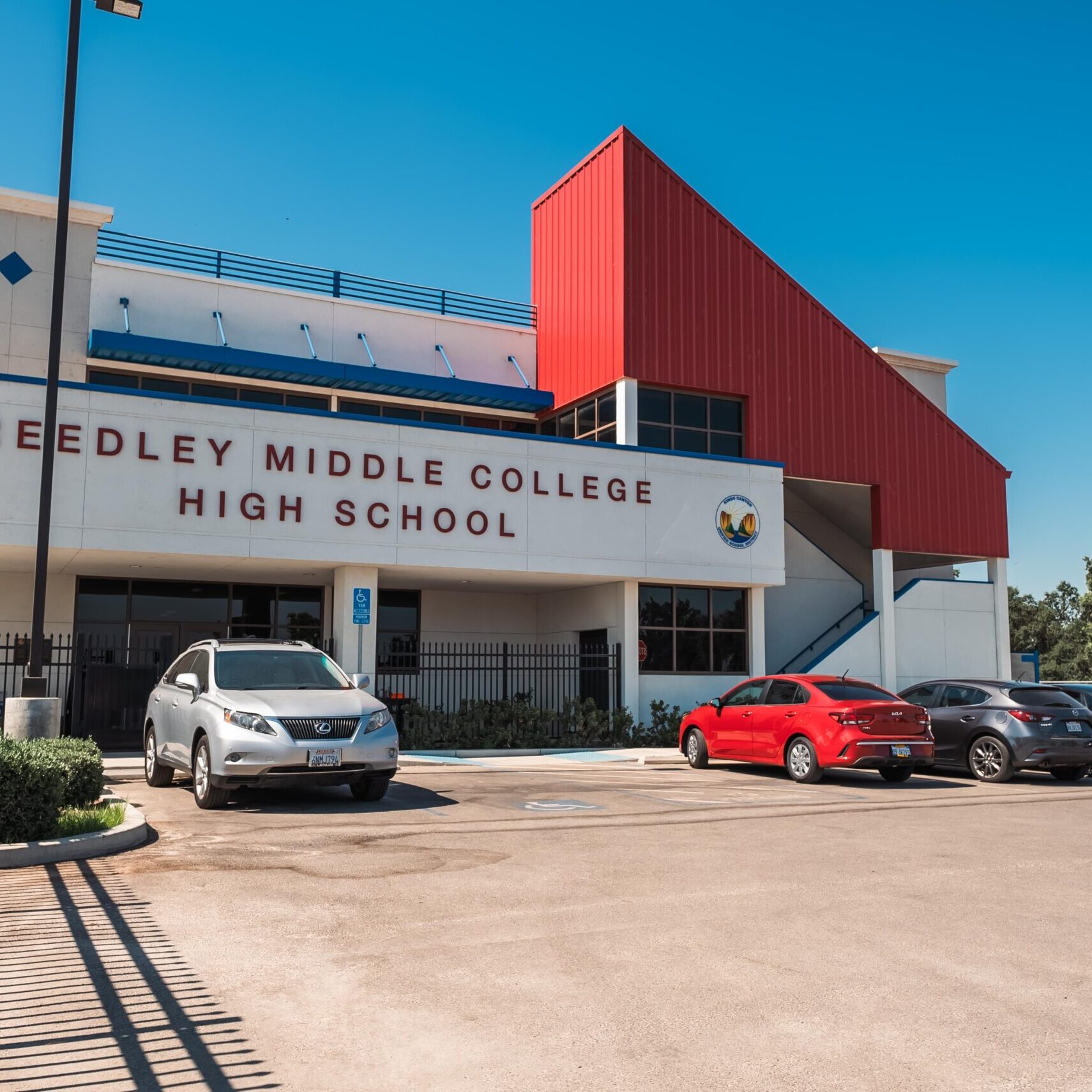 A professional, outdoor photo of the main entrance to Reedley Middle College High School. The modern building has a white facade with blue trim and a striking red metal-paneled roof section. The name "REEDLEY MIDDLE COLLEGE HIGH SCHOOL" is prominently displayed on the front. A black metal fence runs along the building, and a paved parking lot with several cars is in the foreground. The sky is a clear, bright blue.