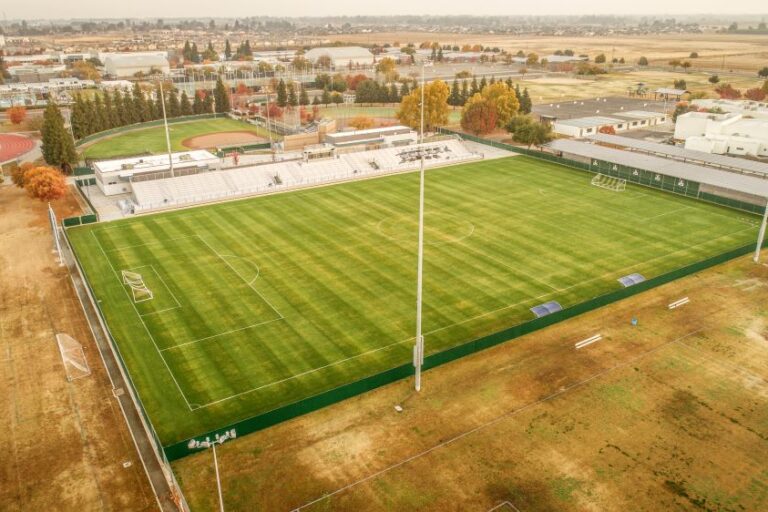 An aerial view of a large, well-maintained sports field, which is the Clovis East Soccer Stadium. The field is covered in bright green grass, with white lines marking a soccer pitch. A set of white bleachers sits on one side of the field, and a baseball field is visible behind it. The stadium is surrounded by a brown field, and in the background, a cluster of residential and commercial buildings are visible under a hazy sky. A row of solar panels is visible on the roof of a building adjacent to the field.