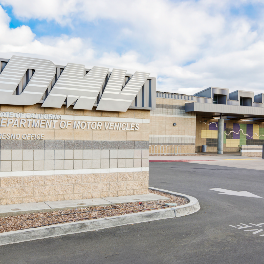 A wide, outdoor, daytime shot of a modern DMV building. The image shows the entrance to the building and a large sign with the stylized, metallic "DMV" logo. Below the logo, the sign reads "STATE OF CALIFORNIA DEPARTMENT OF MOTOR VEHICLES FRESNO OFFICE" in black letters on a light stone block base. The building has a modern, multi-colored facade with light stone and metal panels. The sky is cloudy.
