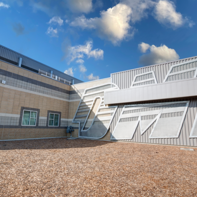 A ground-level, outdoor shot of the side of a modern DMV building. The building has a tan, brick facade and a gray, metallic facade with various sizes and shapes of windows. A large, stylized "DMV" is sculpted into the facade. The foreground is a patch of brown mulch or wood chips, and the sky is blue with fluffy white clouds.