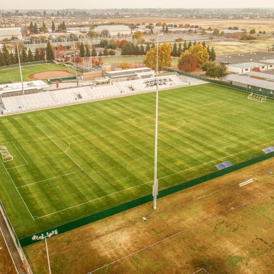 An aerial view of a large, well-maintained sports field, which is the Clovis East Soccer Stadium. The field is covered in bright green grass, with white lines marking a soccer pitch. A set of white bleachers sits on one side of the field, and a baseball field is visible behind it. The stadium is surrounded by a brown field, and in the background, a cluster of residential and commercial buildings are visible under a hazy sky. A row of solar panels is visible on the roof of a building adjacent to the field.