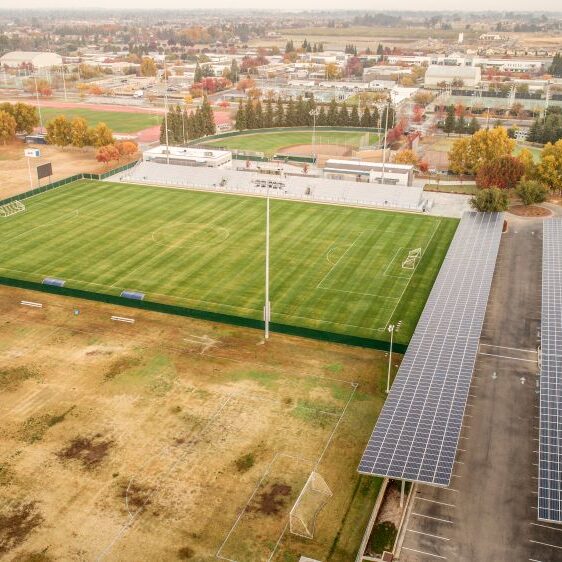 An aerial shot of a large high school sports complex. The image shows a well-maintained soccer field with a set of white bleachers and a press box. Next to the soccer field is a large parking area covered with solar panels. In the background, there is a baseball diamond and a running track. Trees with fall foliage are scattered throughout the campus, and a residential neighborhood is visible in the distance. The sky is a hazy gray.