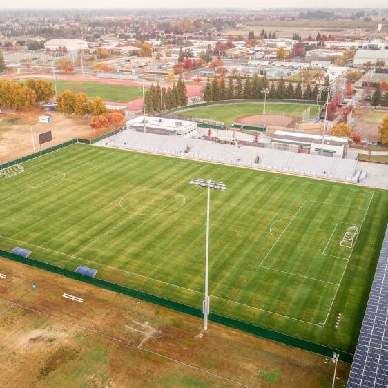 An aerial shot of a large high school sports complex. The image shows a bright green soccer field in the foreground, with white lines and goals. To the right of the field is a large array of solar panels over a paved lot, and on the opposite side are white bleachers and a press box. In the background, a running track, baseball diamond, and other fields are visible, with trees showing fall colors and a residential neighborhood in the distance. The sky is hazy and overcast.