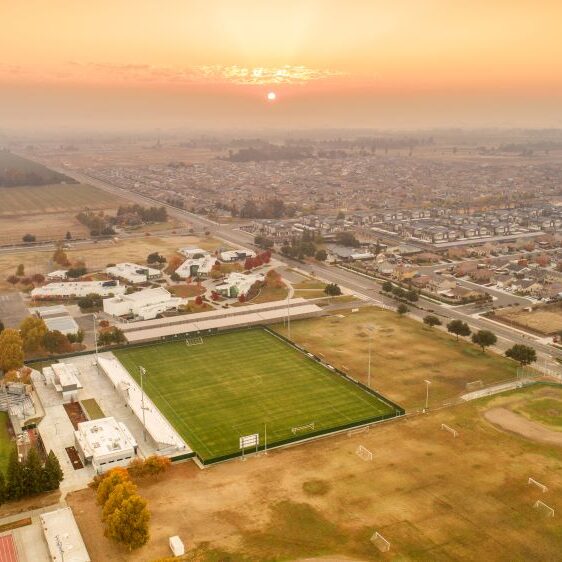 An aerial shot of a large high school sports complex and a surrounding residential area at sunset. In the foreground, there are multiple athletic fields, including a baseball diamond and a soccer stadium. A row of residential homes lines the street to the right. The sky is hazy and orange, and the sun is visible in the middle of the shot, setting over the distant buildings and fields.