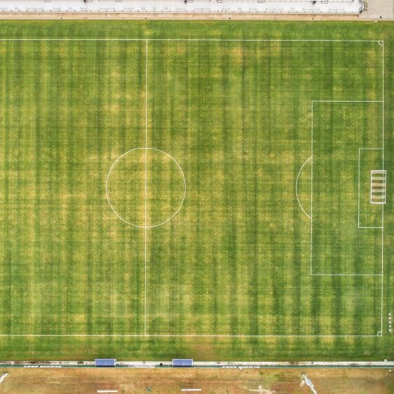 An aerial, top-down view of a soccer field with white lines painted on the grass. The field is in the center, with a large, covered parking area with solar panels on the right. Another, less-maintained field is at the bottom, and a fence is visible along the left side. The goalposts are at either end of the field.