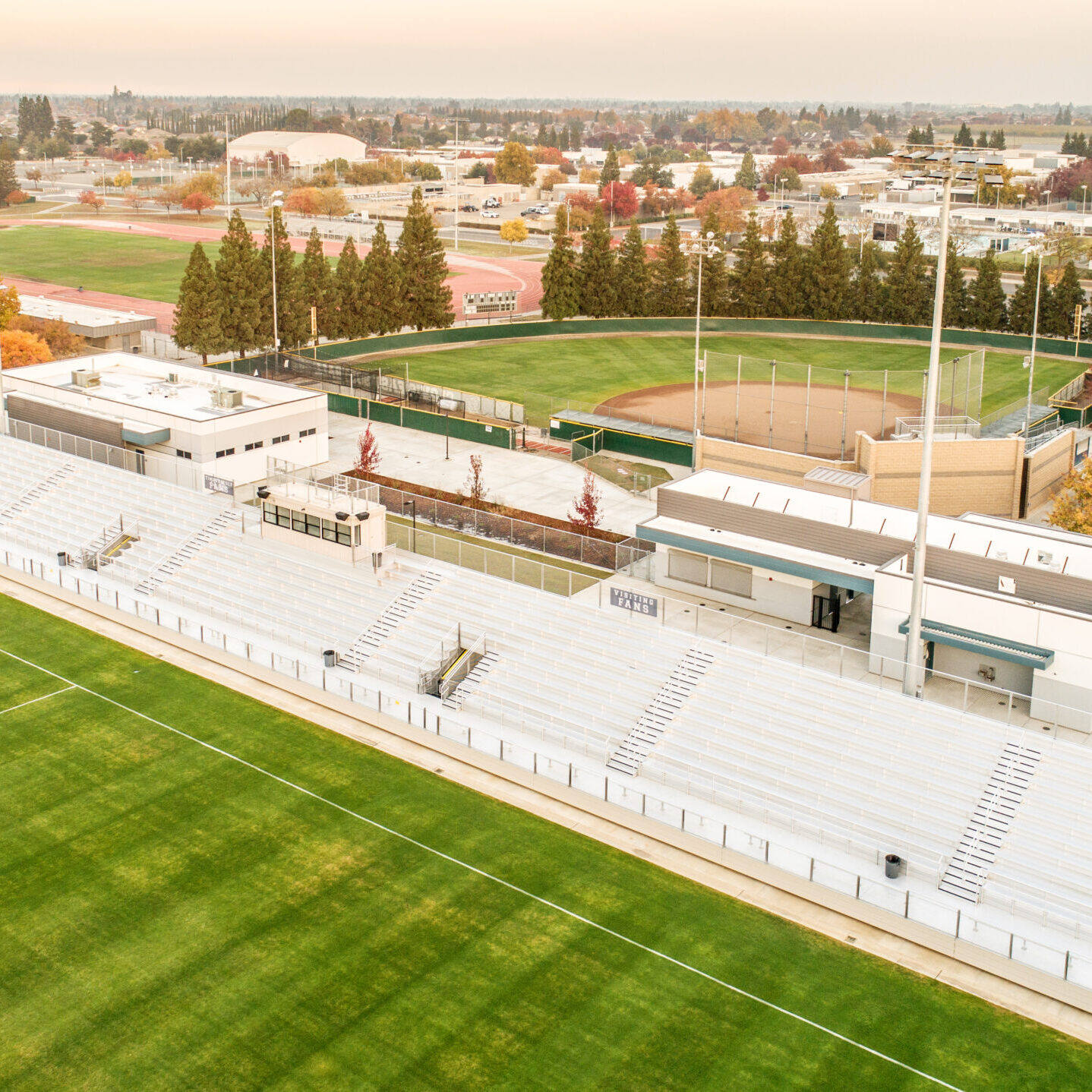 An aerial, eye-level shot of a high school sports complex. The photo shows a large, green soccer field on the left and a set of white bleachers and a press box in the foreground. A baseball field is visible beyond the bleachers. The complex is surrounded by trees with fall colors, and in the distance, a large residential neighborhood and commercial buildings are visible under a hazy sky.