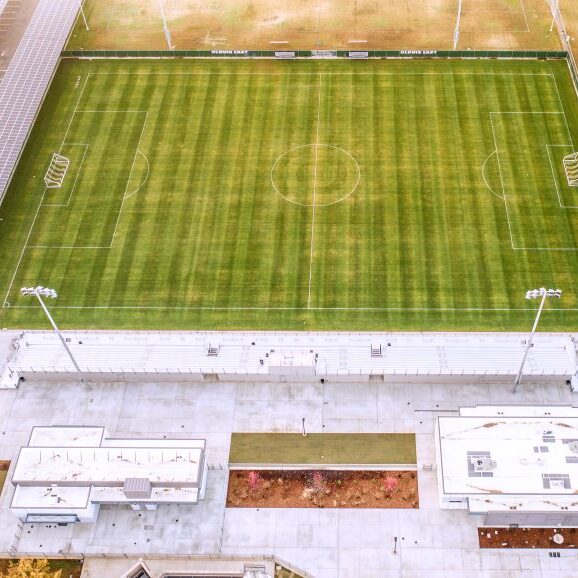 An aerial, top-down view of a soccer stadium at Clovis East High School. The photo shows the bright green soccer field with white markings, surrounded by a green fence with the words "CLOVIS EAST" visible on the far side. Below the field are white bleachers, a press box, and two white buildings. A large array of solar panels covers a parking area or walkway to the left of the field. The ground is a mix of paved concrete and brown earth, with a few trees showing fall colors.