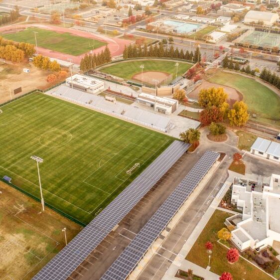 An aerial view of a large high school campus sports complex. The photo shows a well-maintained soccer field with a set of white bleachers and a press box. Next to it is a baseball field, and in the background are a running track and tennis courts. A long row of solar panels covers a walkway and parking area in the foreground. The surrounding area includes other school buildings and residential neighborhoods, with trees showing fall colors. The lighting is hazy, suggesting late afternoon.
