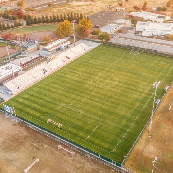 An aerial shot of a large high school sports complex. The image shows a well-maintained soccer field with a set of white bleachers and a press box. Next to the soccer field is a baseball diamond. A long row of solar panels is visible on a covered walkway behind the soccer field. The surrounding area includes other athletic fields and buildings, with residential neighborhoods and trees with fall foliage in the distance. The overall lighting is warm, indicating sunset.
