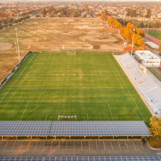 An aerial shot of a large, well-maintained soccer stadium and a series of practice fields at Clovis East High School. A large green soccer field with white markings is in the center, with white bleachers and a press box to the right. The foreground is a paved parking lot with a large array of solar panels, and a line of trees with fall foliage is visible. The surrounding area includes more soccer fields, a running track, and residential rooftops in the distance. The sky is hazy and the light is soft, indicating it is late in the day.