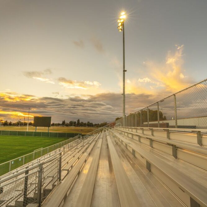 A ground-level shot of a soccer stadium's bleachers at sunset. The image is taken from the side of the stadium, showing the long, empty rows of metal benches, which are reflecting the warm light of the setting sun. A large floodlight pole with a bright light on top is in the foreground, and a chain-link fence runs along the top of the bleachers. The soccer field is visible to the left, and the sky is a mix of gold, orange, and gray clouds.