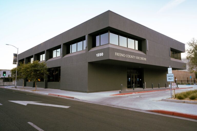 A wide-angle, outdoor shot of a modern, two-story commercial building, which is the Fresno County Recorder's office. The building is a dark gray color with large, dark-tinted windows. The address, "1250", and the name "FRESNO COUNTY RECORDER" are visible on the facade. The photo is taken from a street corner, showing the building's main entrance and a sidewalk with a street sign that says "ENTER ONLY." The sky is a light blue, and a tree is visible on the left.