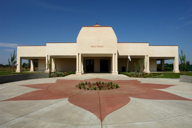 A symmetrical, ground-level exterior shot of a modern Hindu temple building. The facade is a light-colored stucco with a large, elevated central section that has the words "HINDU TEMPLE" on it. A covered portico with a series of pillars extends from the main building. The foreground features a decorative red concrete star or flower pattern embedded in the gray concrete walkway. A small, landscaped garden is in the center of the star pattern. The sky is a clear blue.