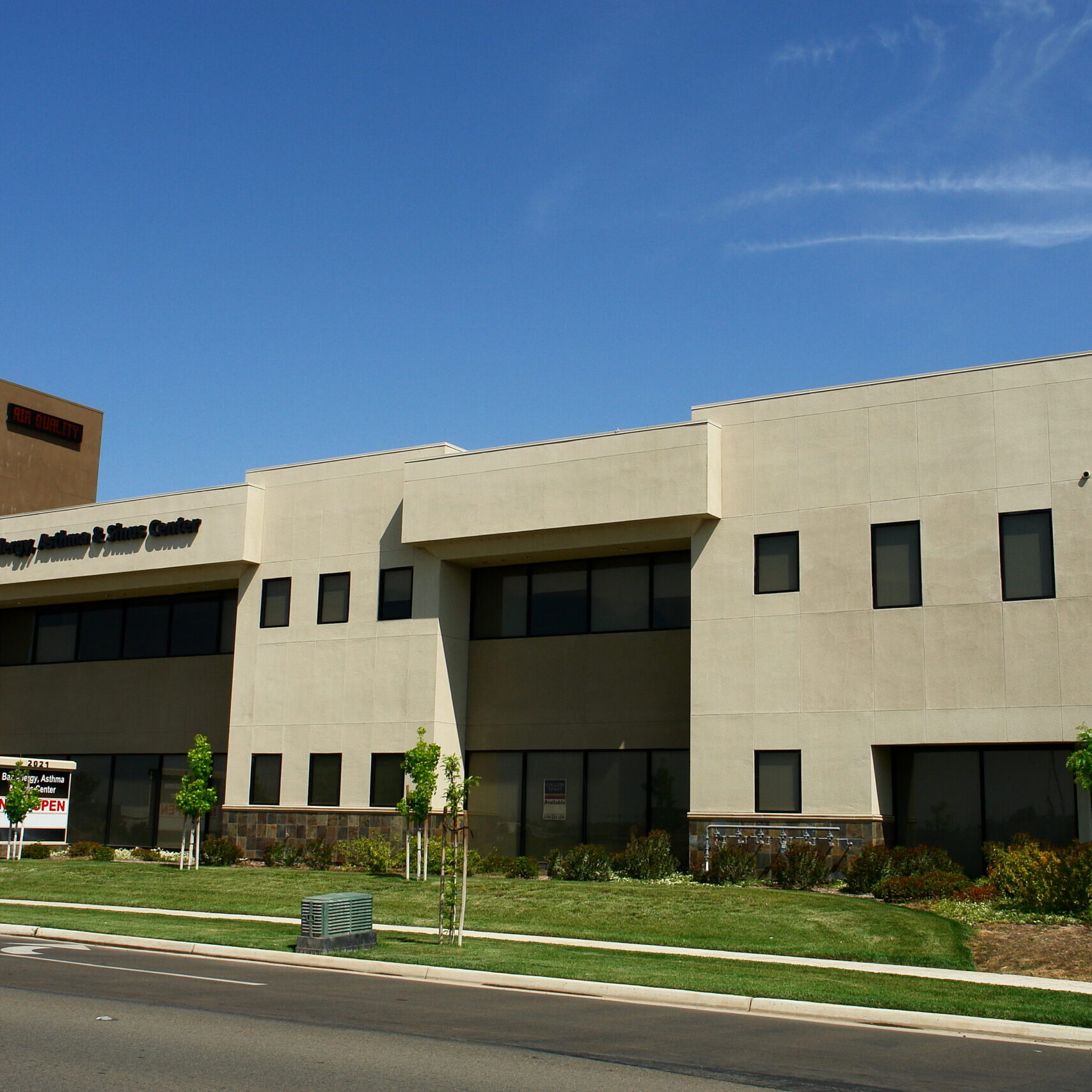 Baz Allergy, Asthma & Sinus Center A full-length exterior shot of a modern, two-story building on a sunny day. The building is made of light-colored brick with large, dark-tinted windows. The left side of the building features a single glass door. The right side has multiple doors and windows. The ground is a mix of a concrete sidewalk and dirt. The sky is bright and clear, and a mountain range is visible in the distant background. The image is taken from a perspective that captures the entire length of the building.