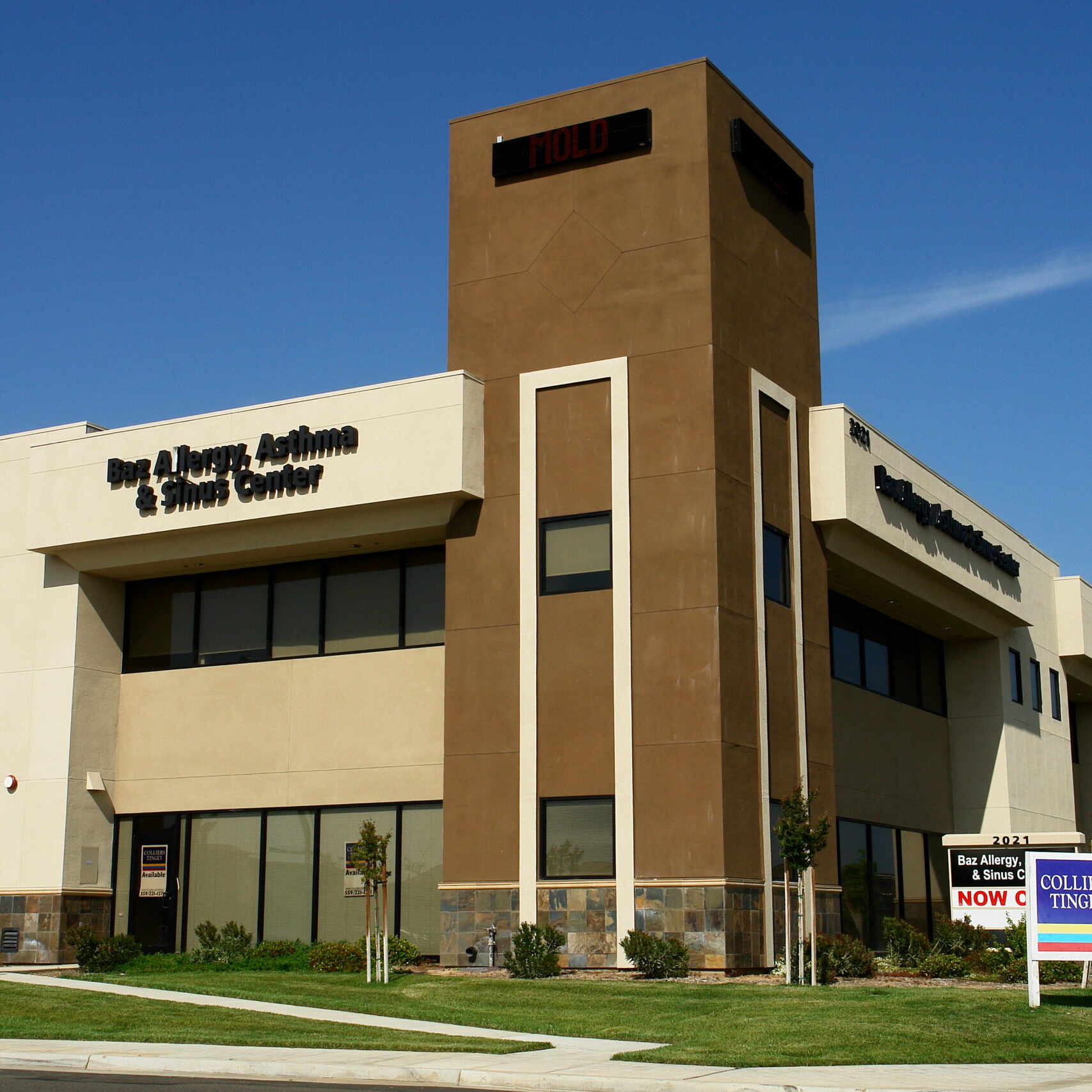 A slightly low-angle, exterior shot of a modern, two-story commercial building on a sunny day. The building's facade is a mix of light-colored stucco, a darker tan central tower, and a stone veneer base. The words "Ear, Allergy, & Sinus Center" are visible on the left side of the building. In the foreground, there is a manicured lawn and two "For Lease/Sale" signs for Colliers Tingey. The sky is a clear, deep blue. The image captures the building from a street-level perspective, highlighting its clean lines and professional appearance.