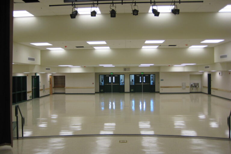 A symmetrical, interior shot of a large, empty multi-purpose room at Oakhurst Elementary School. The room has a highly polished, light-colored floor that reflects the ceiling lights and the double doors at the far end. The walls are a light tan color with a wood trim. Above, the ceiling is a mix of acoustic tiles with square fluorescent lights and a large set of theater spotlights. There are two sets of double doors with "EXIT" signs above them on the back wall. The room appears to be ready for an event or activity.