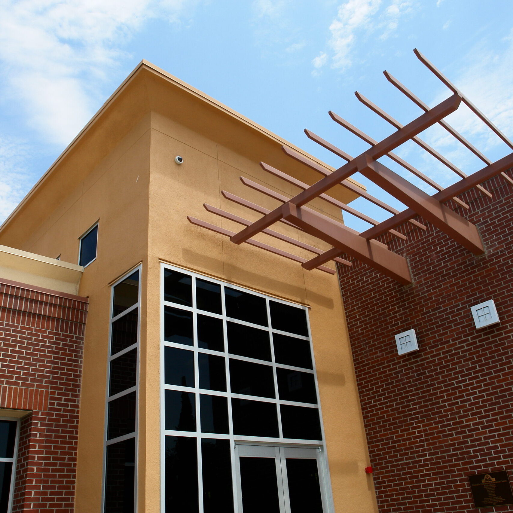An eye-level exterior shot of a modern, two-story building on a sunny day. The building is made of red brick and light tan stucco. A section of the front facade is a large window with a white grid and a silver frame. The building has a decorative wooden pergola structure over the entrance. The sky is bright blue with scattered clouds. The image is taken from a low angle, highlighting the different textures and architectural features of the building.