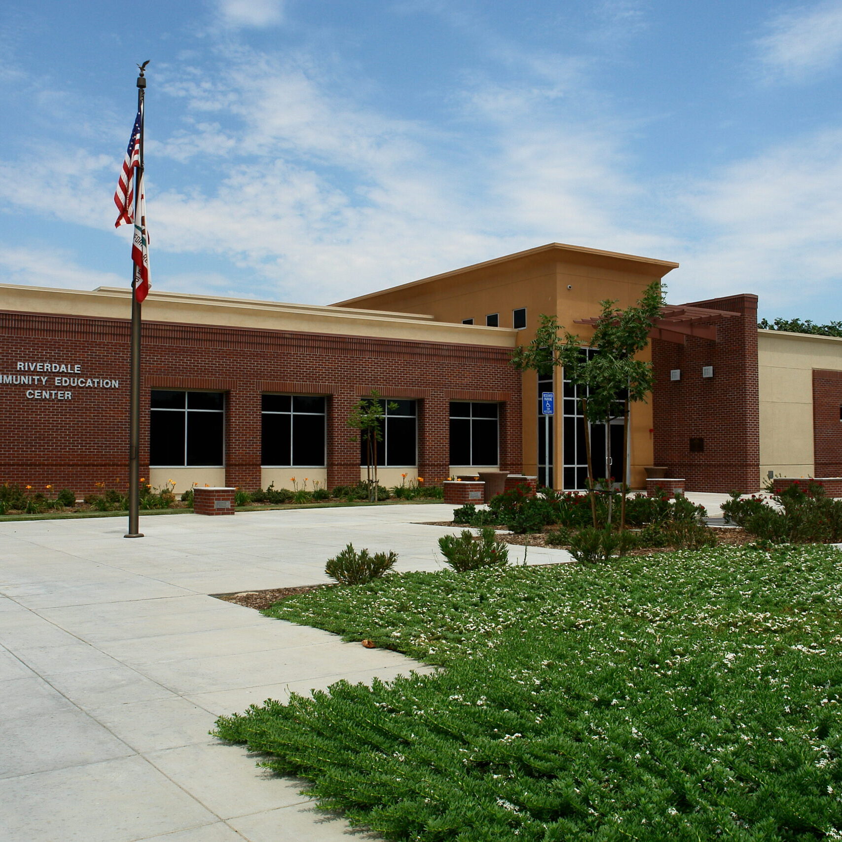 A full-length, wide-angle exterior shot of a modern, one-story community education center on a sunny day. The building has a brown brick facade with a tan-colored stucco section. The words "RIVERDALE COMMUNITY EDUCATION CENTER" and the building number "3160" are visible on the brick wall. A large flagpole with an American flag and a state flag is in front of the building. The foreground has a landscaped area with green grass, shrubs, and a paved walkway. The sky is bright blue with scattered white clouds. The image conveys a clean, professional, and well-maintained public building.