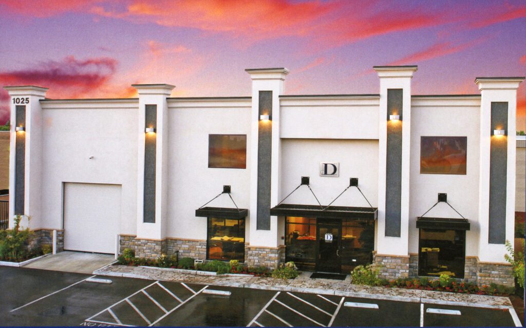 A slightly low-angle, exterior shot of a modern, one-story commercial building at dusk. The building has a light-colored stucco facade with dark gray accent panels and a stone veneer base. Three prominent columns with decorative caps frame the front. A large glass entrance with a dark awning is in the center, and two smaller windows with awnings flank it. A large garage door is on the left side of the building. The building number "1025" and a letter "D" are visible on the facade. The sky is filled with a dramatic mix of orange, pink, and purple clouds.