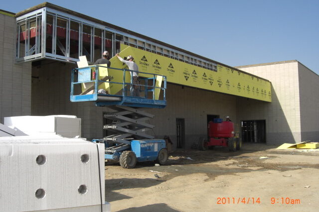 A daytime construction photograph shows two workers in a blue scissor lift, applying yellow sheathing to the exterior of a building. The sheathing is branded with the logos of "DensGlass" and "Georgia-Pacific". The building's walls are made of concrete masonry units (CMU), and a steel-framed canopy or overhang extends from the structure. A red lift is visible in the background, and construction materials are scattered on the dirt ground in the foreground, including a pallet of what appears to be concrete blocks. The sky is bright and clear. A timestamp at the bottom right corner reads "2011/4/14 9:10am".