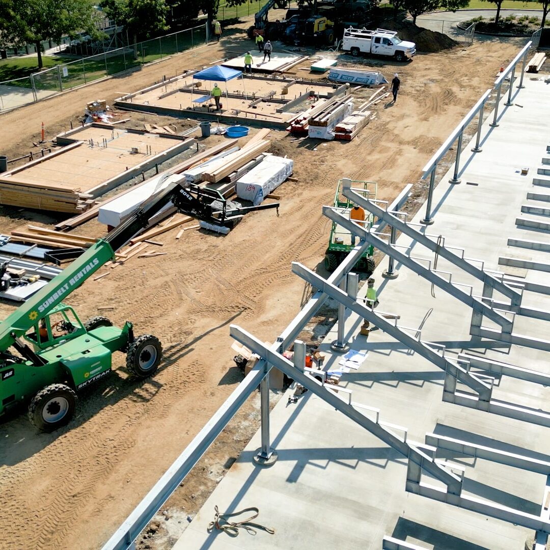 a wide aerial shot of a green forklift setting up a row of beams that will become bleachers on a sunny afternoon with a dirt field behind the construction site