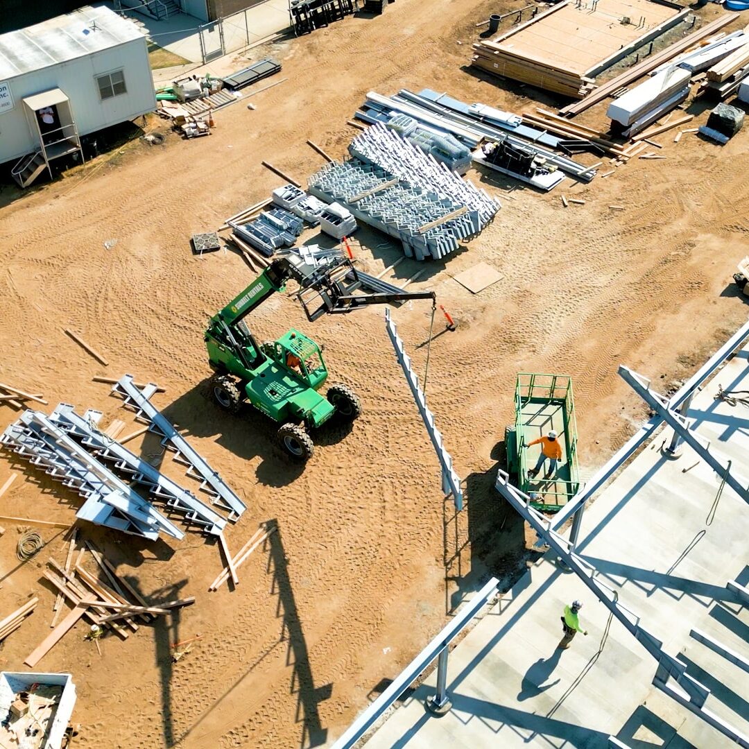 An aerial shot of a dirt field with various construction trucks, forklifts, work trailer, and multiple beams that will be installed with direct sun facing down on the scene
