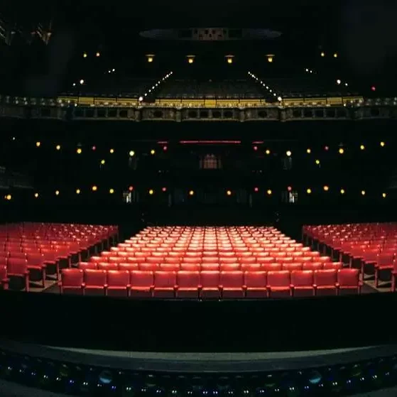 A low-light, wide-angle photograph of the inside of a theater from the stage's perspective. The stage floor curves in the foreground, with an orchestra pit area. Rows of empty red seats fill the auditorium, divided into three main sections. The lighting is dim, with small, golden-glowing lights suspended from the high ceiling, which is not fully visible. The decorative architecture of the theater, possibly in a classical style, is dimly lit in the upper parts of the frame. The overall atmosphere is dark and intimate, with all the focus on the rows of empty chairs.