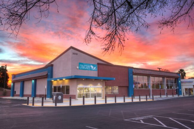 A dramatic, wide-angle exterior shot of the Betty Rodriguez Library building at sunset or sunrise. The sky is filled with vibrant hues of orange, pink, and purple clouds. The one-story library building has a contemporary design, with a light-colored central section and a darker red and blue section to the right. A blue awning with the word "LIBRARY" and an owl logo is above the main entrance. Bare tree branches are visible in the foreground, framing the top of the image. The parking lot in front of the building is empty, with black bollards lining the walkway. The building number "3040" is visible near the roofline.
