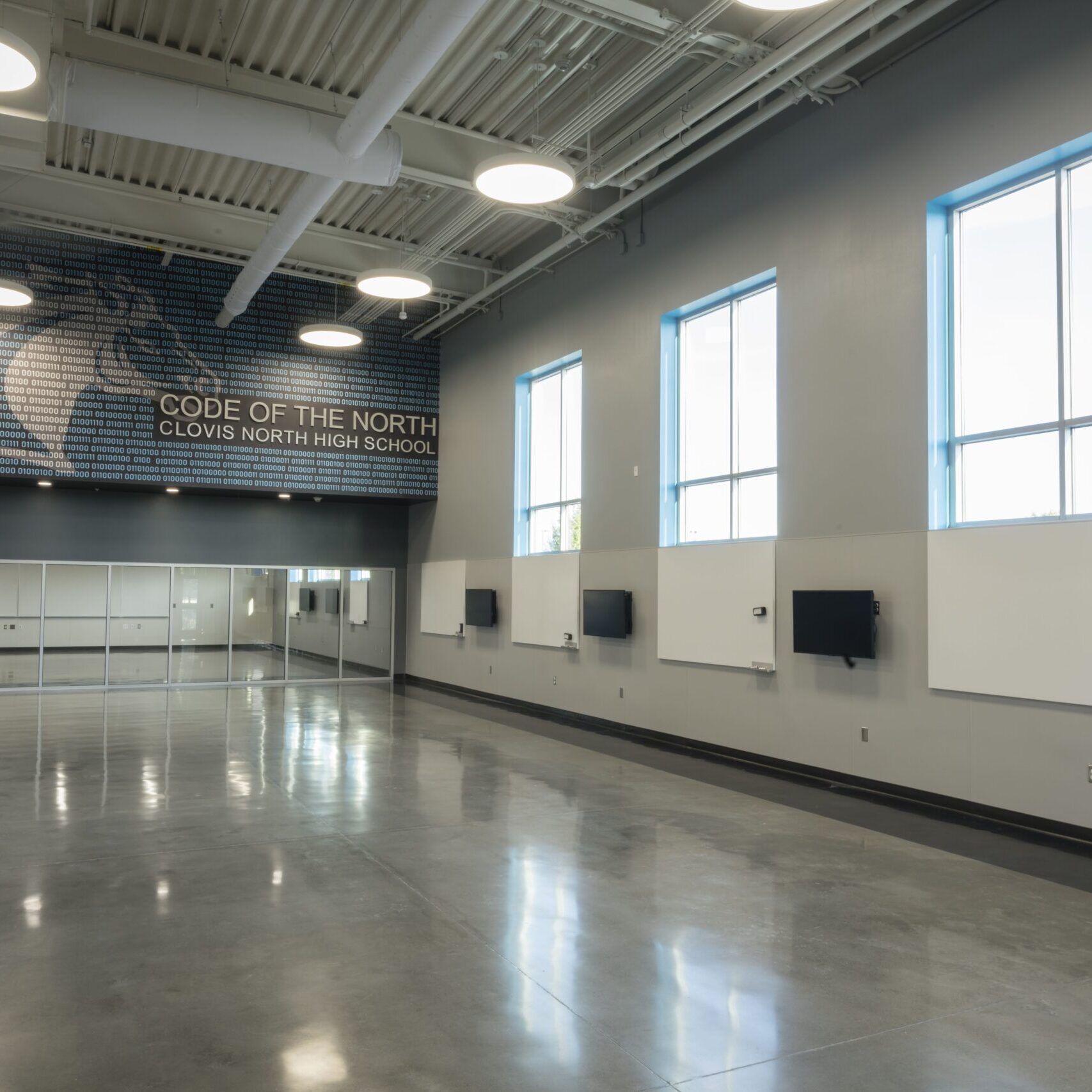 An interior view of a large, modern, empty room with polished concrete floors and high ceilings. A large mural on the far wall says "CODE OF THE NORTH CLOVIS NORTH HIGH SCHOOL." There are large windows on the right and a wall of glass panels on the left.